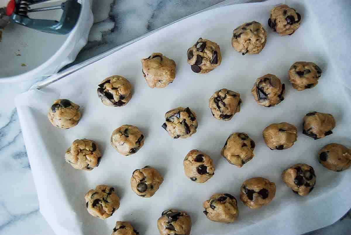 A baking sheet lined with parchment paper holds rows of unbaked chocolate chip cookie dough balls. A cookie scoop and a bowl are partially visible on the side.
