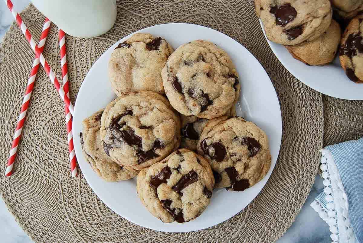 A white plate holds six chocolate chip cookies with large chocolate chunks, next to a glass of milk.