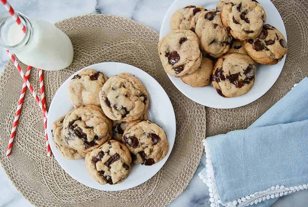 Two white plates filled with browned butter chocolate chip cookies sit on woven placemats next to a bottle of milk with red-striped straws.