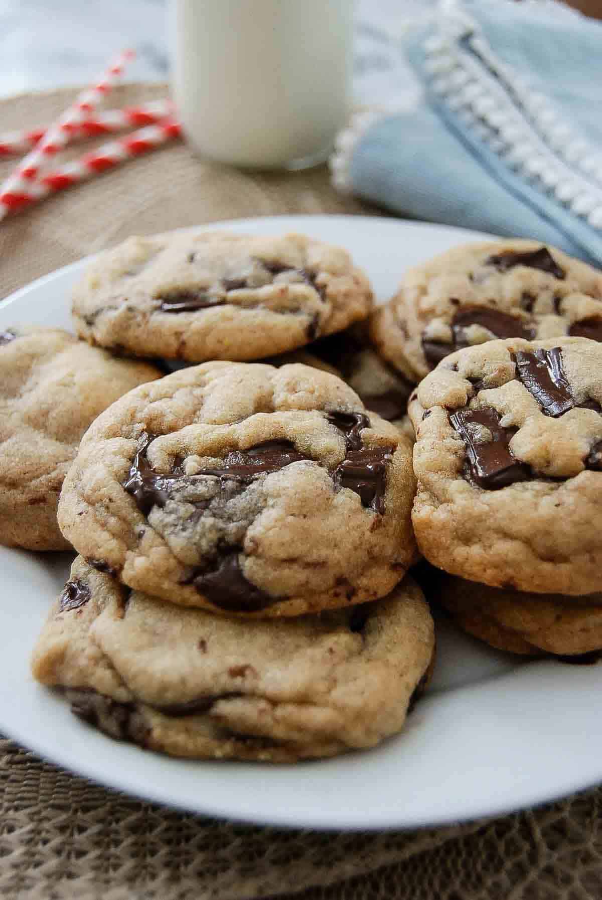 A white plate filled with thick, chewy brown butter chocolate chip cookies sits on a table next to a glass of milk.