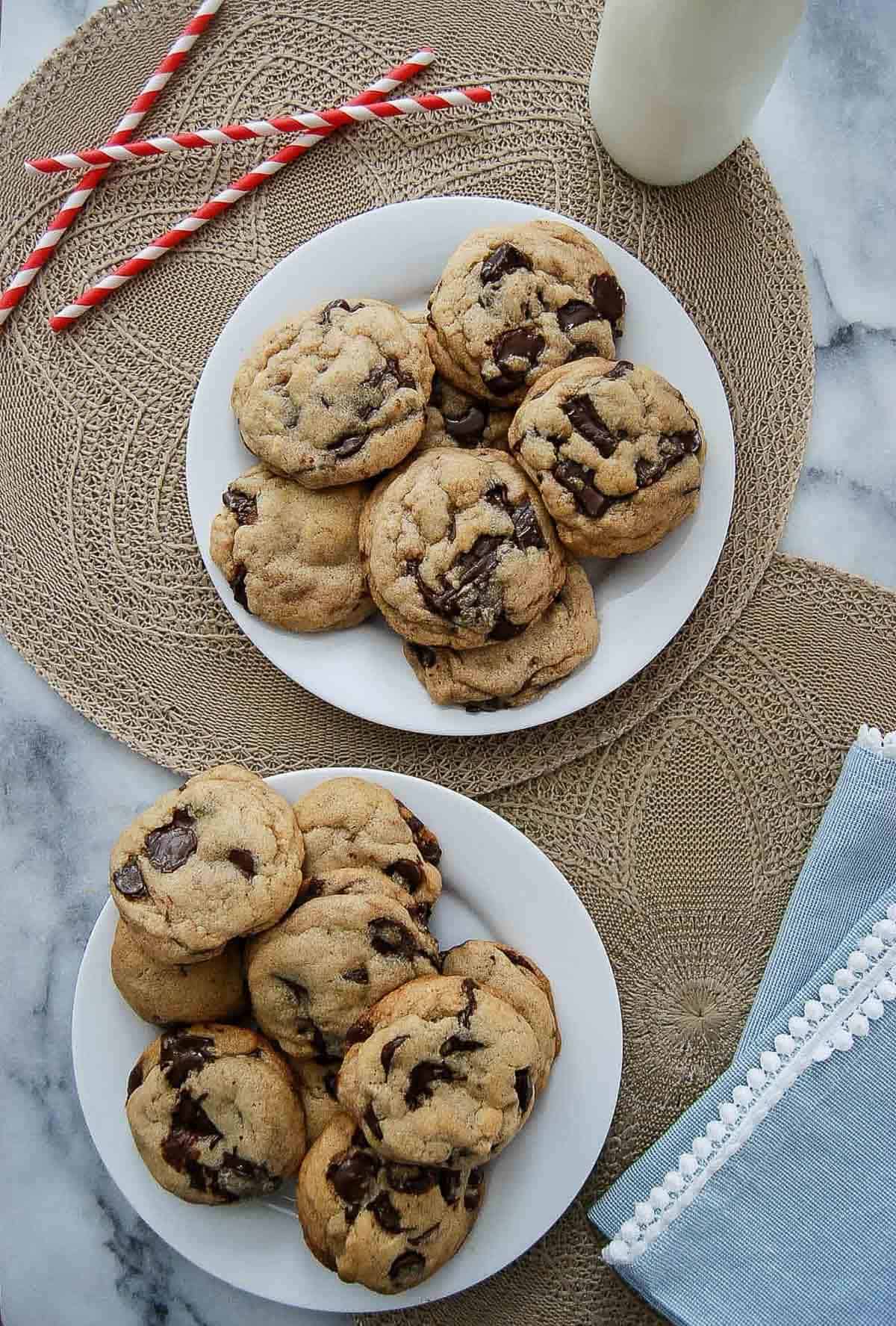 Two white plates filled with brown butter chocolate chip cookies sit on woven placemats, with three red-striped straws and a glass of milk nearby, next to a blue napkin with white trim on a marble surface.