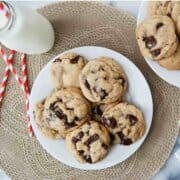 A plate of brown butter chocolate chip cookies sits on a woven placemat, with a glass bottle of milk and red striped straws nearby. More cookies are on a plate in the corner.