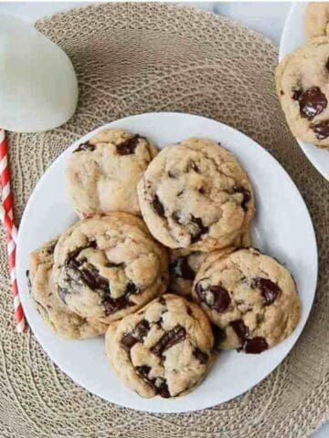 A plate of brown butter chocolate chip cookies sits on a woven placemat, with a glass bottle of milk and red striped straws nearby. More cookies are on a plate in the corner.