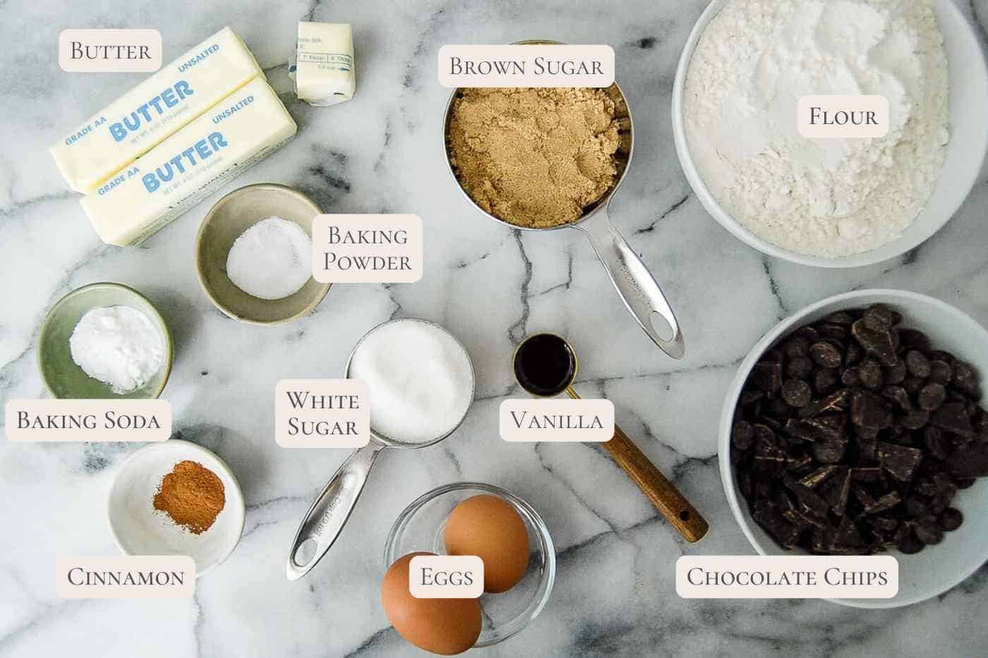 Baking ingredients for brown butter chocolate chip cookies on a marble surface, including butter, brown sugar, flour, baking powder, baking soda, white sugar, vanilla, cinnamon, eggs, and a bowl of chocolate chips, each labeled with text.