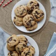Two white plates filled with brown butter chocolate chip cookies on beige placemats, with red striped straws, a bottle of milk, and a blue napkin with white trim on a marble surface.