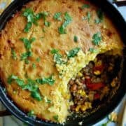 A round baking dish filled with a casserole topped with cornbread, garnished with chopped cilantro. A large wedge is missing, revealing a filling of ground meat, beans, corn, and bell peppers.