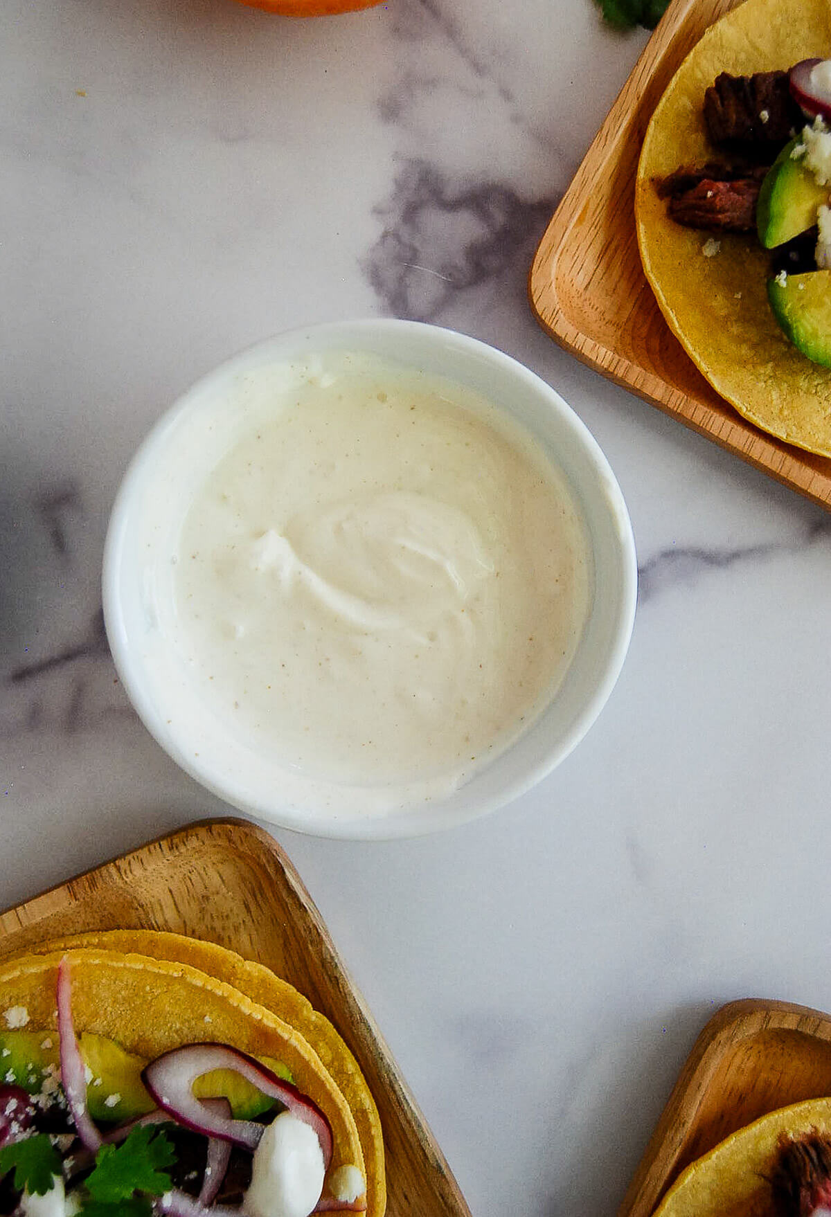 A small white bowl filled with lime crema sits on a marble surface, surrounded by wooden plates holding tacos with various toppings like avocado, cilantro, and onions.
