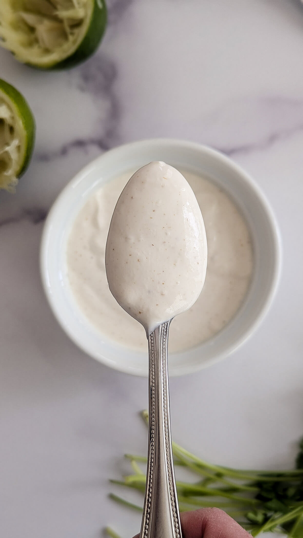 A hand holds a spoonful of lime crema sauce above a small bowl of the same sauce, with squeezed limes and green herbs visible in the background on a marble surface.