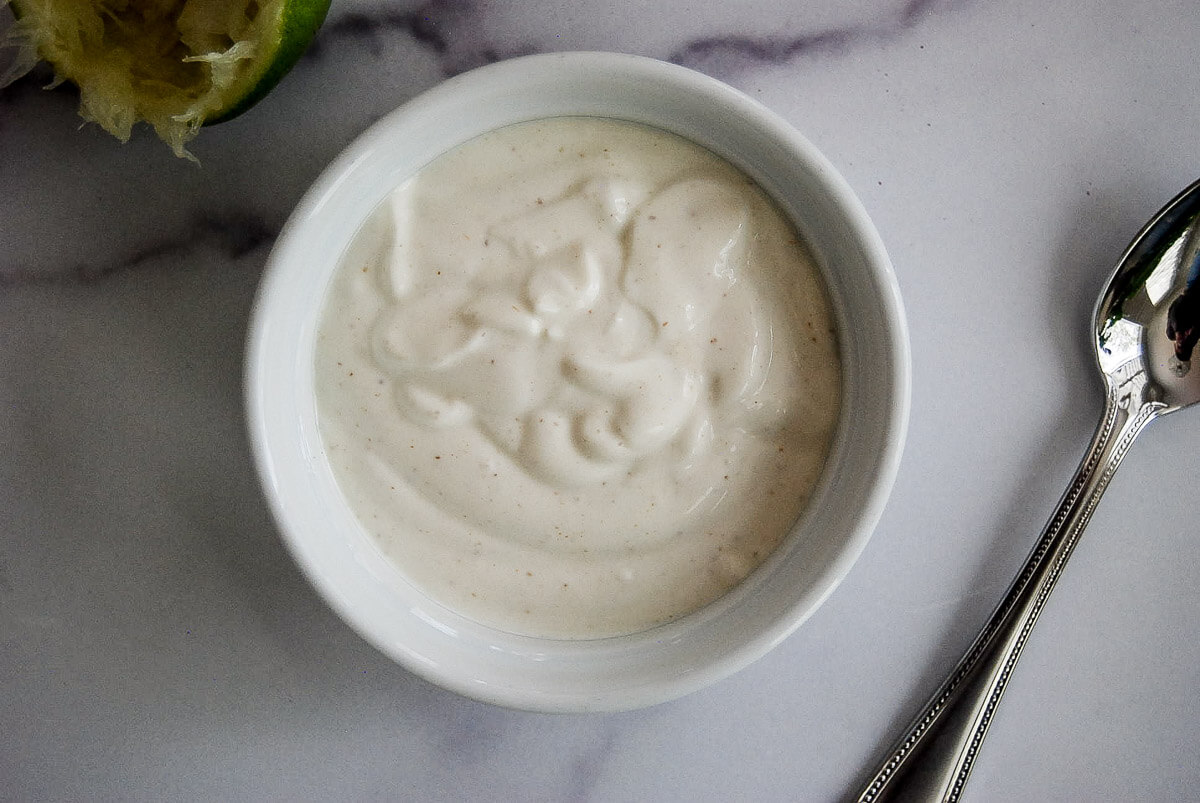 A small white bowl filled with lime crema sits on a marble surface next to a spoon and a partially squeezed lime.