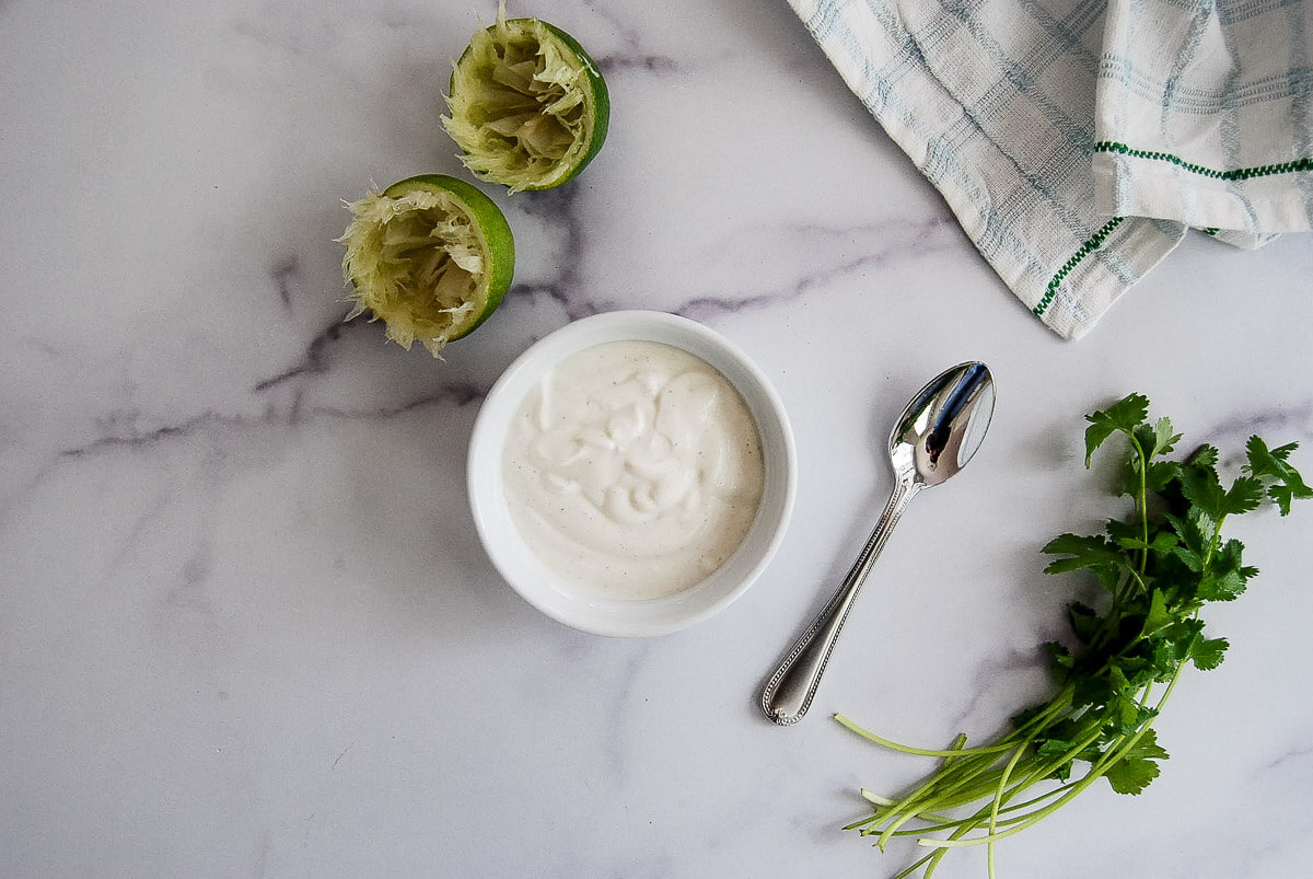 A bowl of lime crema sits on a marble surface next to a spoon, squeezed lime halves, fresh cilantro, and a checkered kitchen towel.