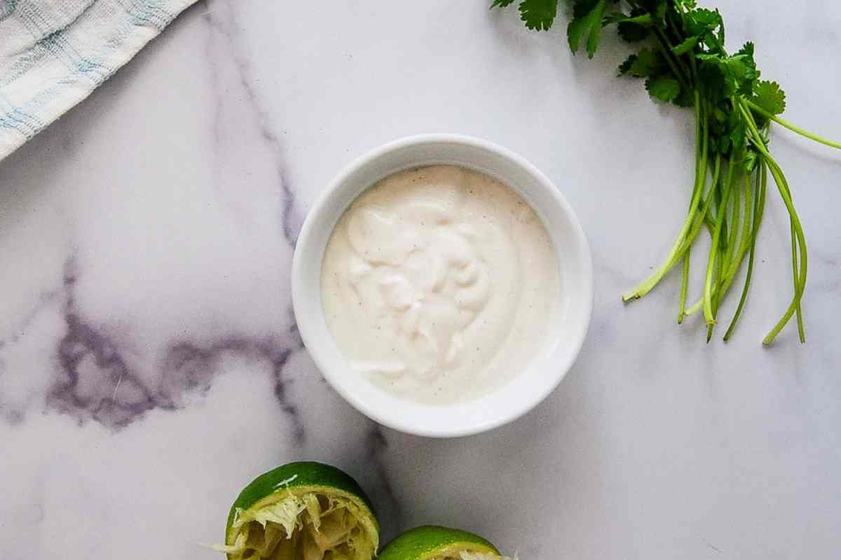 A small white bowl of lime crema sits on a marble surface, next to squeezed lime halves, and sprigs of fresh cilantro.