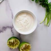 A small white bowl filled with lime crema sits on a marble surface. Beside it are two squeezed lime halves and fresh cilantro sprigs.