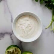 A white bowl filled with lime crema sits on a marble surface beside two squeezed lime halves and a sprig of fresh cilantro.