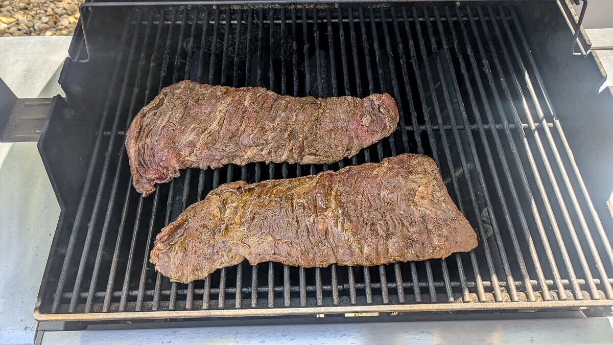 Two pieces of seasoned skirt steak are grilling on an outdoor barbecue, placed side by side on the grill grates over the heat.