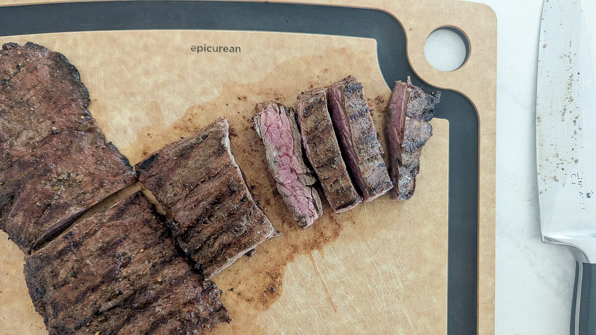 Sliced grilled skirt steak on a brown cutting board with a large knife next to it.