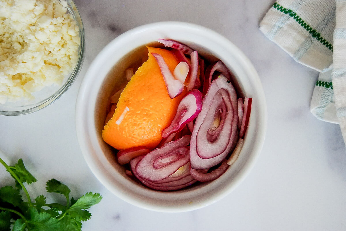 A small white bowl with pickled red onions and an orange peel, next to a bowl of crumbled cheese, a sprig of cilantro, and a striped towel on a white surface.