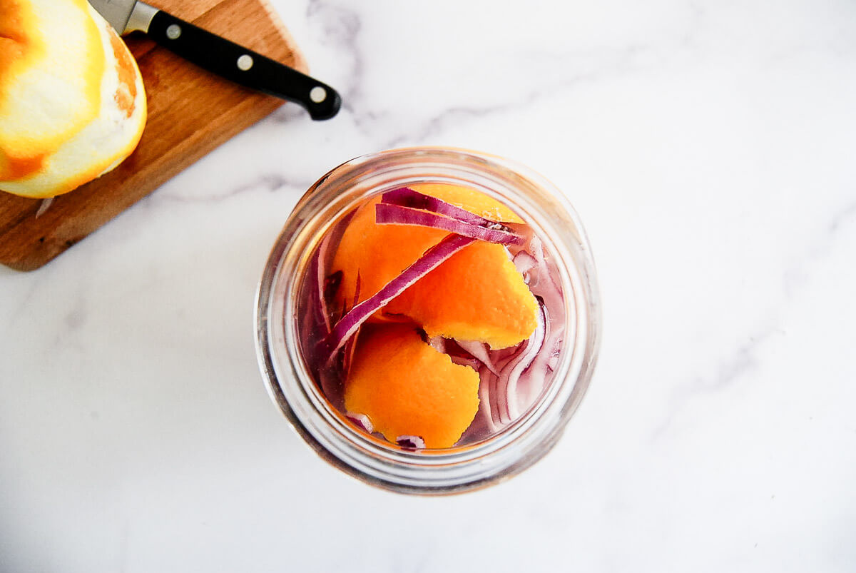 A glass jar filled with peeled orange segments and slices of red onion sits on a white surface; a cutting board with a paring knife nearby.