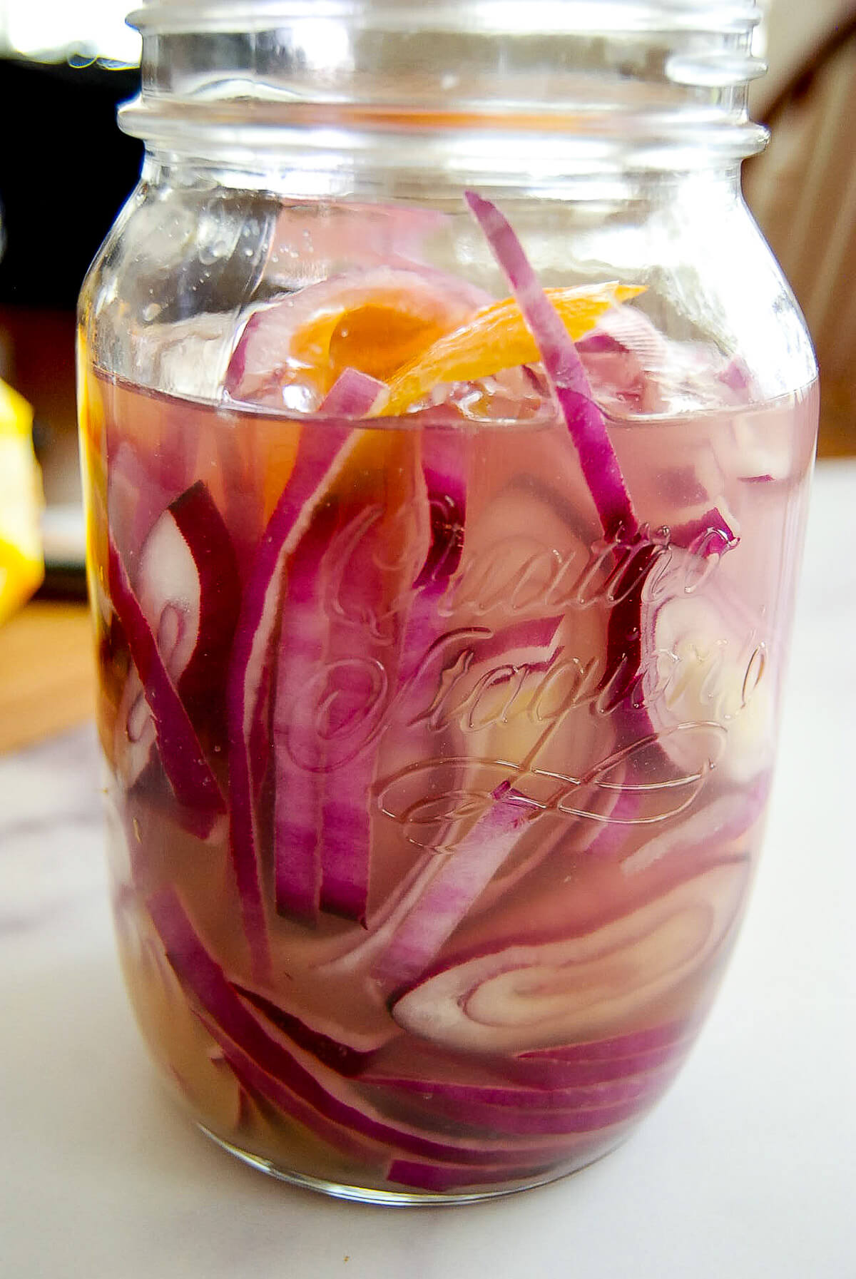 A glass jar filled with pickled red onions and a few orange slices soaking in a clear pickling liquid, sitting on a white surface.