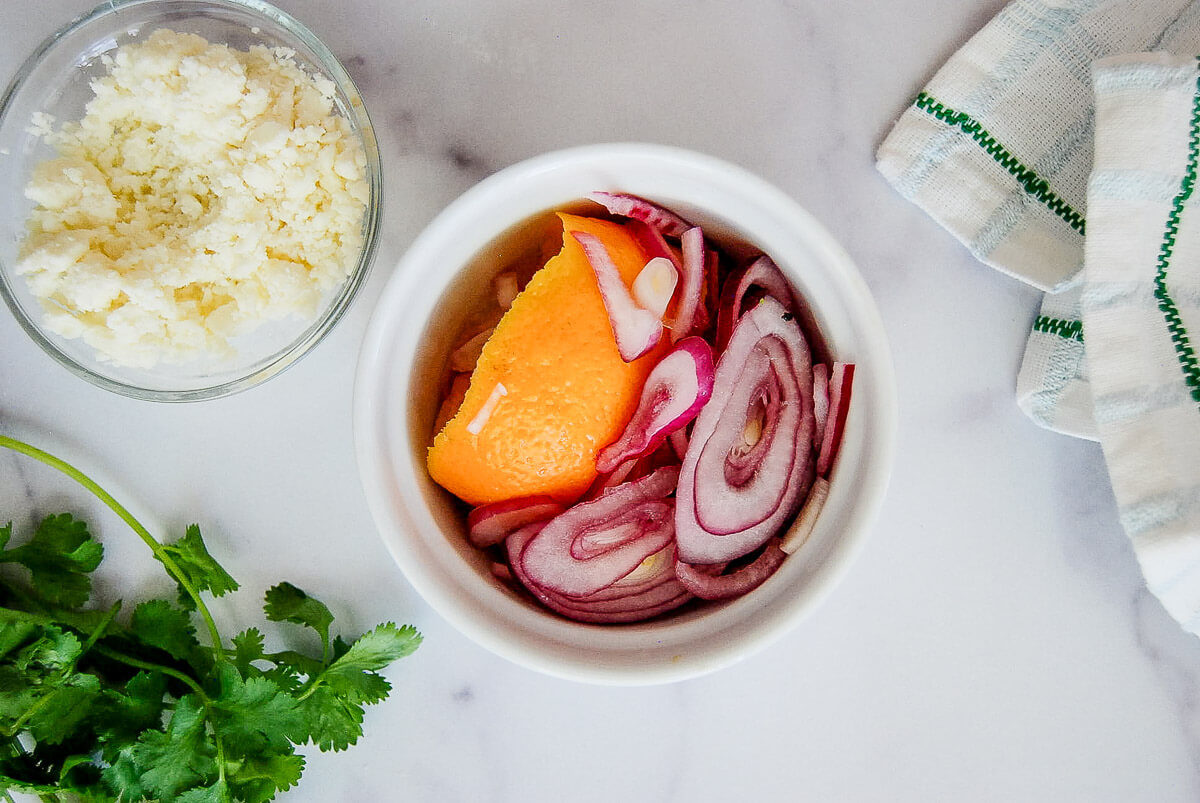 A bowl of pickled red onions and an orange slice next to a bowl of crumbled cheese, fresh cilantro, and a white kitchen towel on a marble countertop.