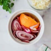 A small white bowl filled with pickled red onions and an orange peel, sitting on a white surface next to cilantro, crumbled cheese in a glass bowl, and a patterned napkin.
