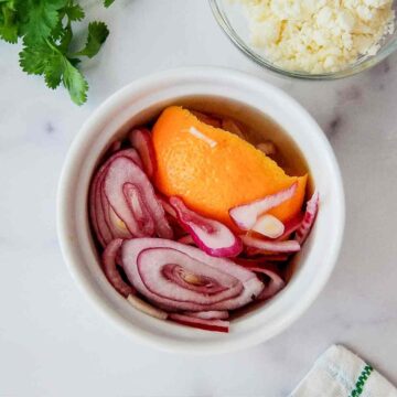 A small white bowl filled with pickled red onions and an orange peel, sitting on a white surface next to cilantro, crumbled cheese in a glass bowl, and a patterned napkin.