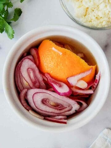 A small white bowl filled with pickled red onions and an orange peel, sitting on a white surface next to cilantro, crumbled cheese in a glass bowl, and a patterned napkin.