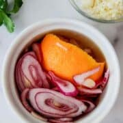 A white bowl filled with pickled red onions and an orange peel, next to a bowl of crumbled white cheese and fresh cilantro on a white surface.