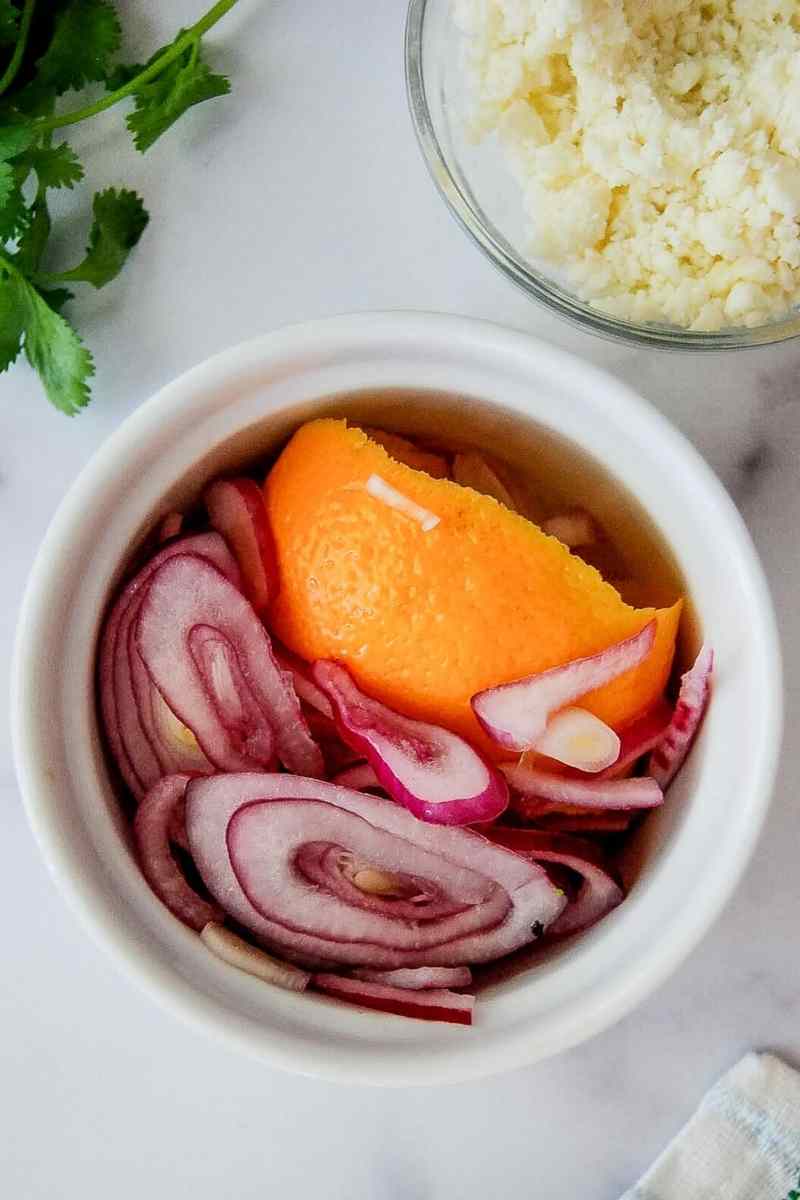 A white bowl filled with pickled red onions and an orange peel, next to a bowl of crumbled white cheese and fresh cilantro on a white surface.
