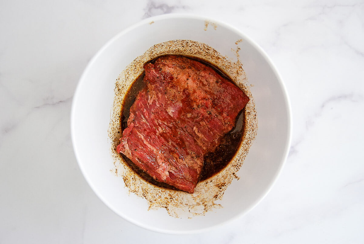 A piece of raw skirt steak is marinating in a skirt steak marinade in a white bowl, placed on a light marble surface.