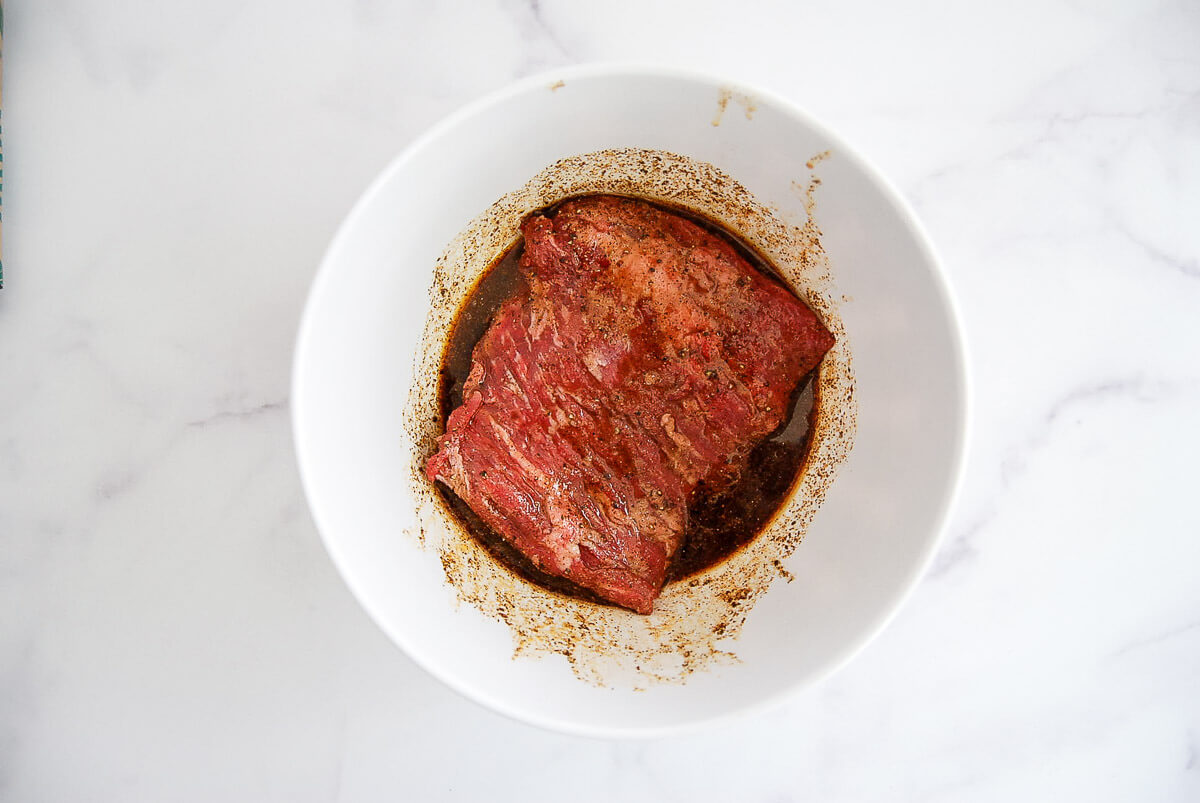 A piece of raw skirt steak is marinating in a white bowl with a dark liquid marinade, set on a white marble surface.