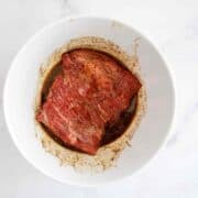 A piece of raw skirt steak marinating in a dark, seasoned liquid inside a white bowl, viewed from above on a light surface.