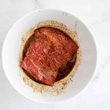 A piece of raw skirt steak marinating in a dark, seasoned liquid inside a white bowl, viewed from above on a light surface.