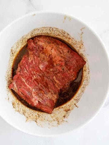 A piece of raw skirt steak marinating in a dark, seasoned liquid inside a white bowl, viewed from above on a light surface.