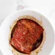 Raw skirt steak marinating in a bowl with dark sauce and spices, set on a white marble surface.