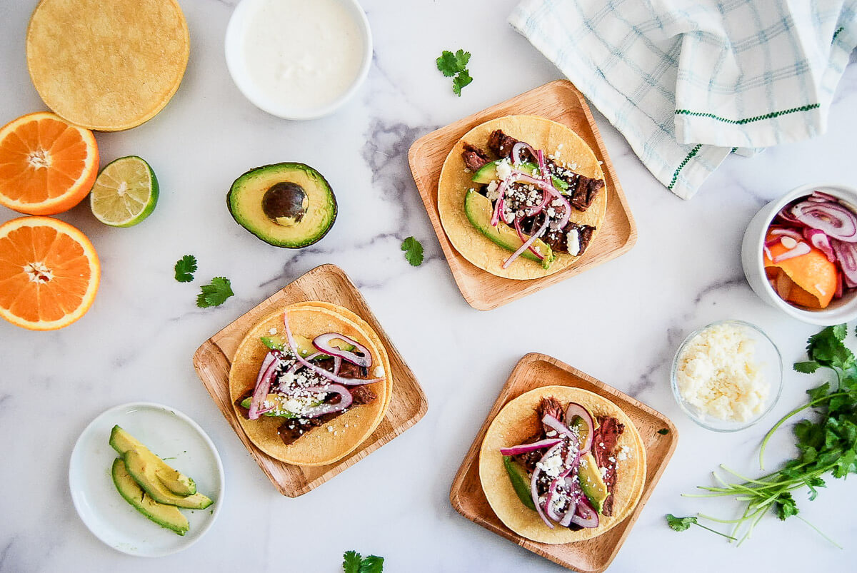 Three small skirt steak tacos topped on wooden plates. Surrounding them are sliced avocado, citrus fruits, cilantro, cheese, and lime crema sauce.