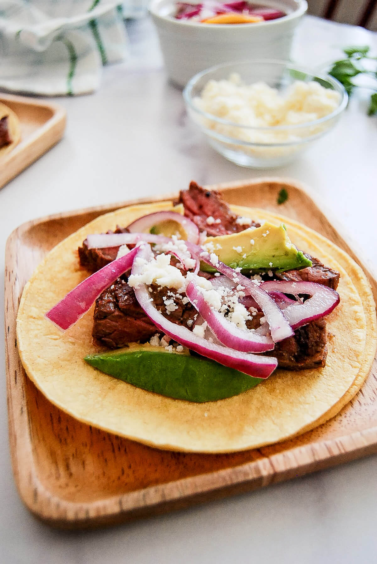 A corn tortilla topped with grilled skirt steak slices, avocado, pickled red onions, and crumbled cheese sits on a wooden plate. Bowls of extra toppings are blurred in the background.