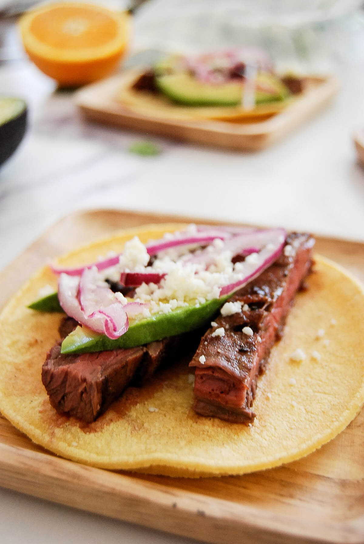 A corn tortilla topped with grilled skirt steak strips, avocado slices, pickled red onions, and crumbled cheese sits on a wooden plate, with another partially visible taco and orange half in the background.
