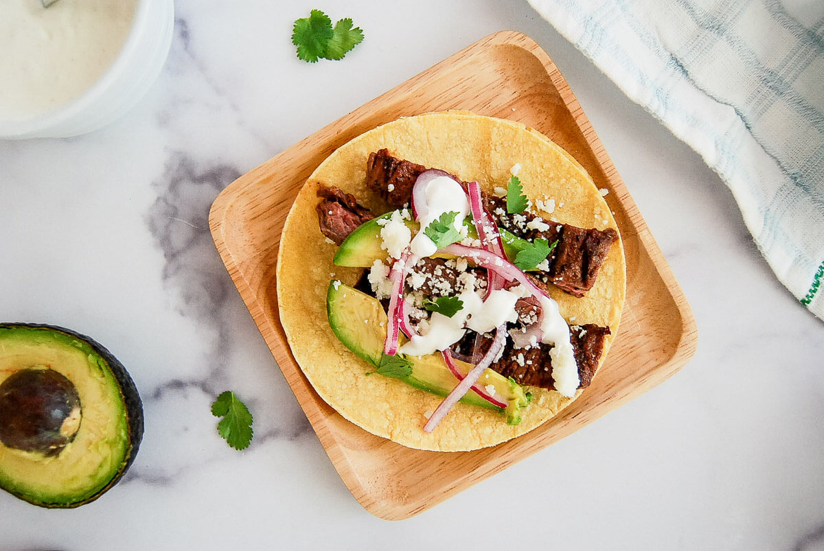 A skirt steak taco on a wooden plate topped with avocado slices, pickled onions, cilantro, cheese, and lime crema. An avocado half, cilantro leaves, and a striped towel are nearby on a white surface.