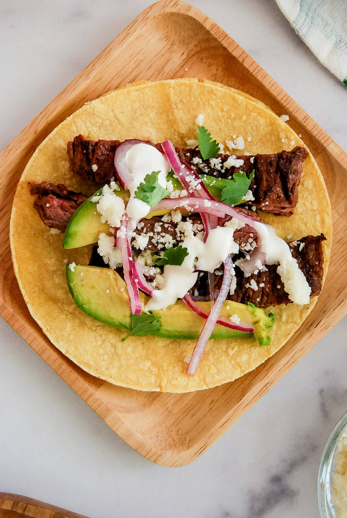 A corn tortilla topped with grilled steak strips, avocado slices, pickled red onions, crumbled cheese, fresh cilantro, and a drizzle of lime crema sauce, served on a wooden plate.