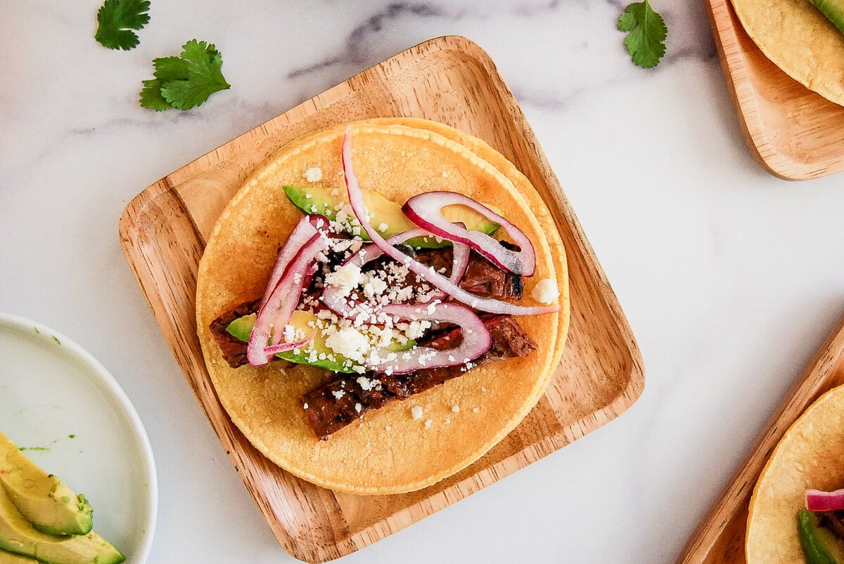 A skirt steak taco on a wooden plate topped with sliced avocado, pickled red onions, and crumbled cheese, garnished with cilantro leaves on a white surface.
