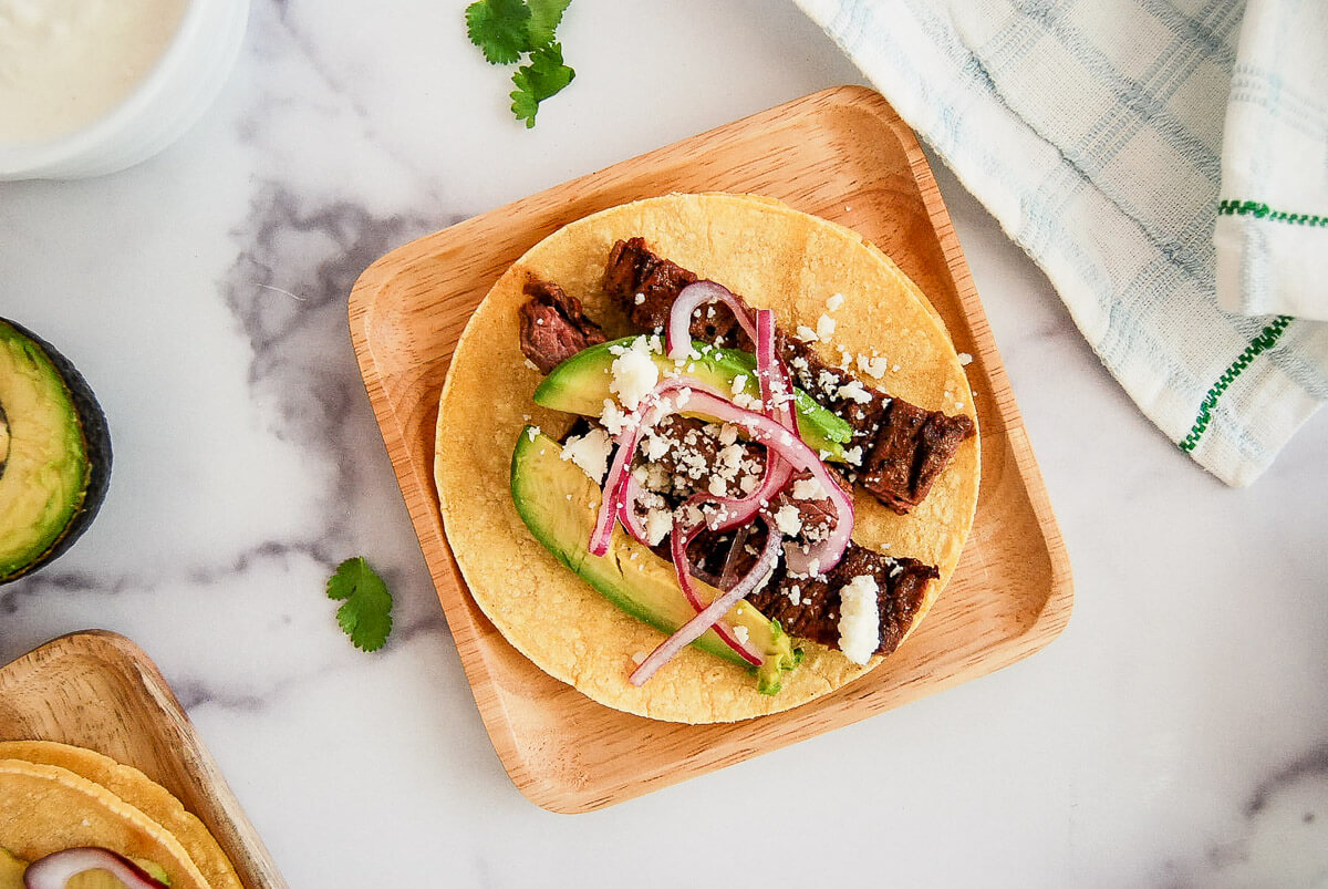 A wooden plate with a taco filled with grilled skirt steak, avocado slices, pickled red onions, and crumbled cheese on a corn tortilla, placed on a marble surface next to a cloth napkin.