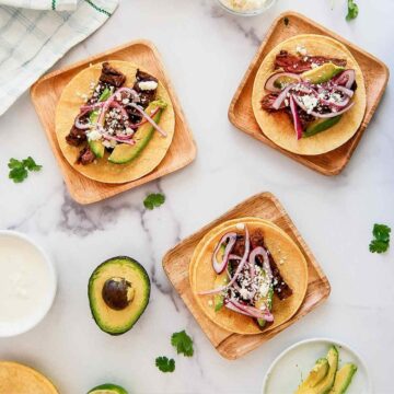 Three wooden plates with skirt steak tacos topped with avocado slices, pickled red onions, and crumbled cheese. Fresh avocado halves, cilantro, and lime are scattered on a white marble surface.
