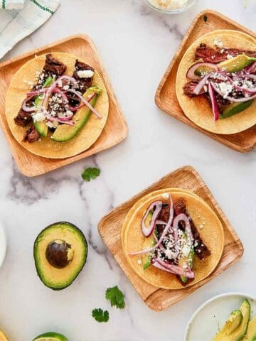 Three wooden plates with skirt steak tacos topped with avocado slices, pickled red onions, and crumbled cheese. Fresh avocado halves, cilantro, and lime are scattered on a white marble surface.
