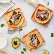 Three small tacos with sliced steak, avocado, pickled red onions, and cheese on wooden plates.