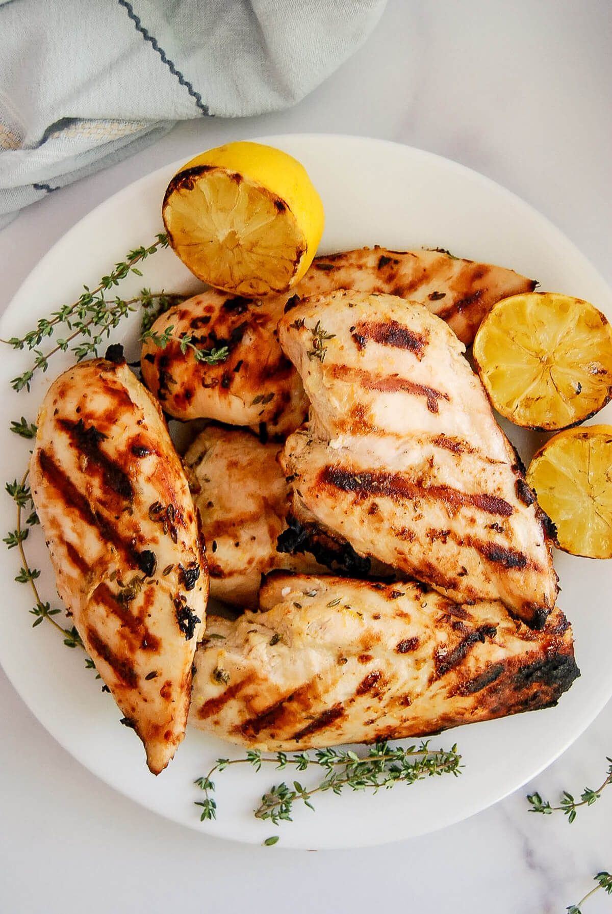 Grilled lemon chicken breasts on a white plate, garnished with fresh thyme and halved grilled lemons, with a light cloth napkin in the background.