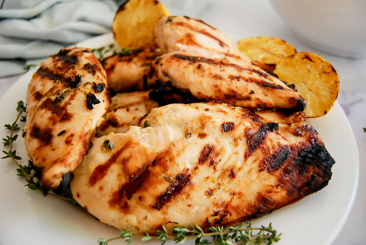 Grilled chicken breasts with char marks on a white plate, garnished with thyme sprigs and grilled lemon halves.