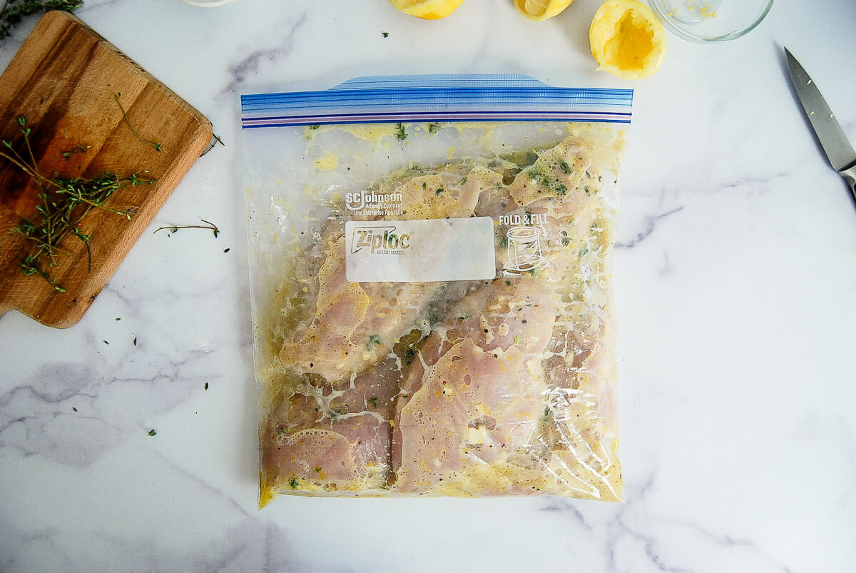 Raw chicken breasts marinating in a sealed Ziplock bag with herbs and lemon on a marble surface. A wooden board with herbs, a cut lemon, and a knife are nearby.