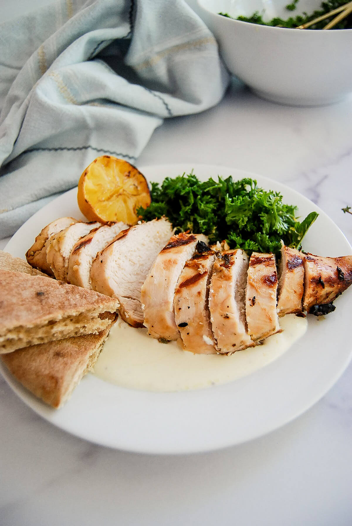 Sliced grilled chicken breast on a plate with pita bread, fresh parsley, a grilled lemon half, and whipped feta, set on a white table with a light blue napkin.