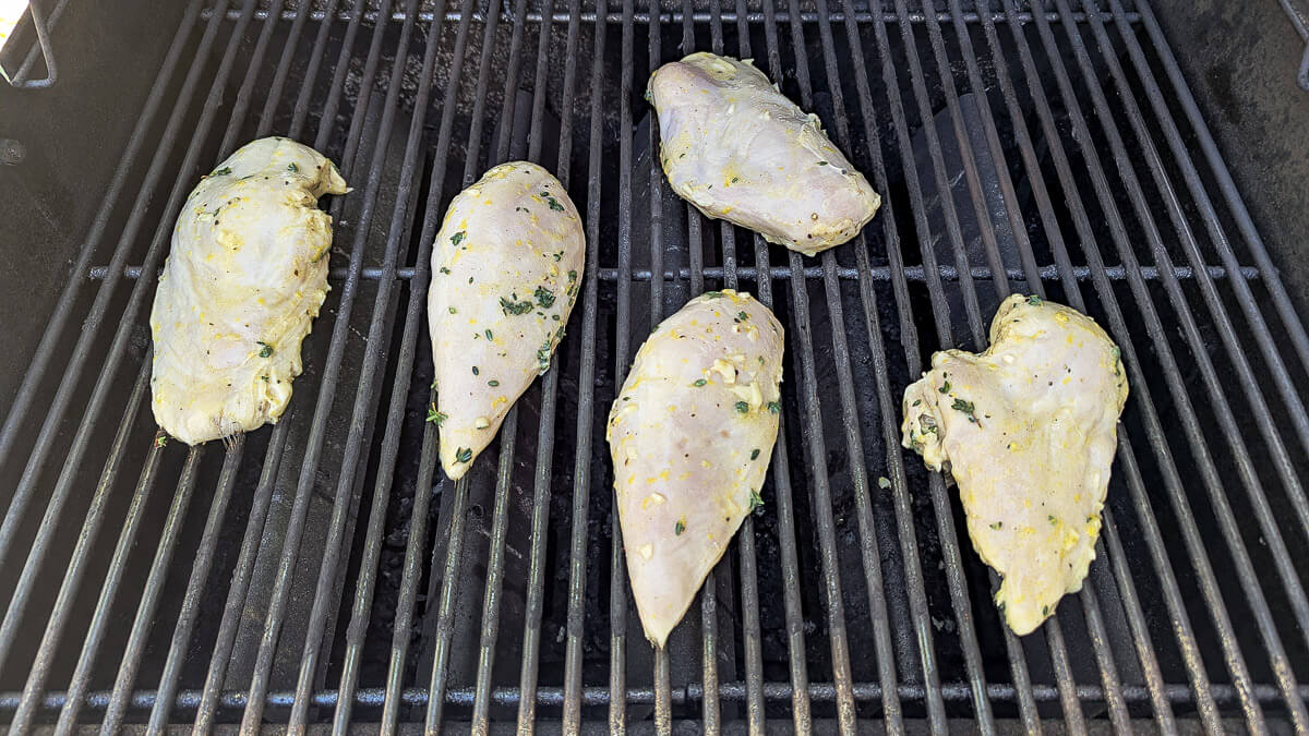 Five marinated chicken breasts with herbs and lemon are placed on a hot grill, spaced apart on metal grates, ready to be cooked.