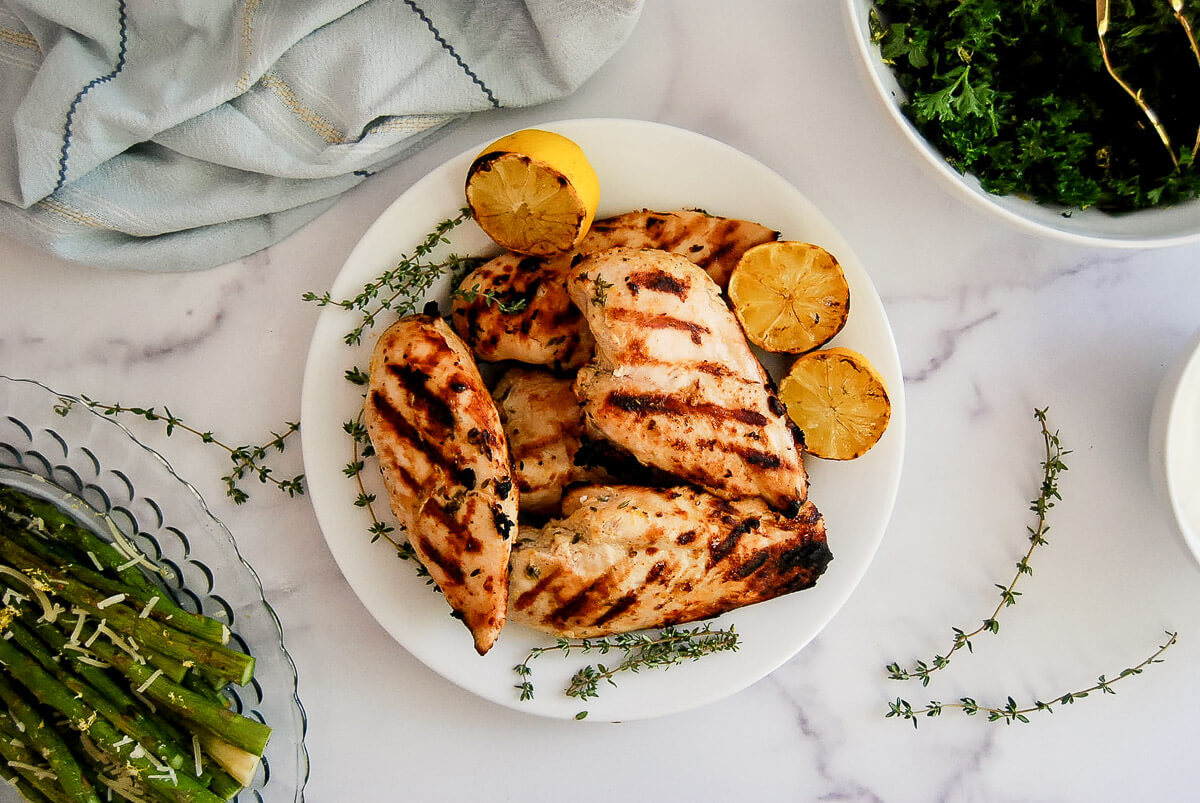 Grilled lemon chicken breasts on a white plate with grilled lemon halves and sprigs of thyme. Asparagus with Parmesan and a bowl of greens are nearby on a white marble surface.